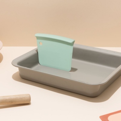 Tabuleiro de forno cinzento com espátula verde dentro, sobre superfície bege