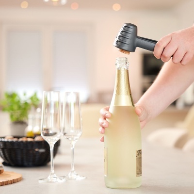 Mãos a abrir garrafa de vinho espumante com abridor preto, com duas taças e ambiente doméstico