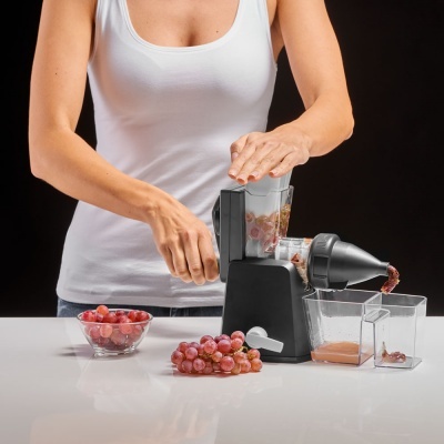 Máquina manual preta para fazer sumo com uvas vermelhas e recipientes transparentes sobre uma superfície branca.