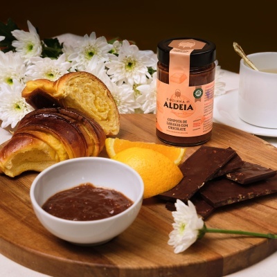 Pequeno-almoço com compota Aldeia, croissant, chocolate, laranja e chá, disposto numa tábua de madeira com flores brancas ao fundo