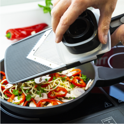 Mandolina de cozinha preta com lâmina em V a fatiar legumes frescos sobre frigideira preta no fogão