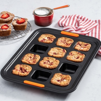 Tabuleiro de forno preto com bolos com morango, coador vermelho e pano de riscas vermelhas