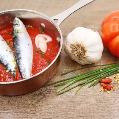 Panela com sardinhas em molho de tomate, alho, tomate, cebolinha e piri-piri sobre mesa de madeira