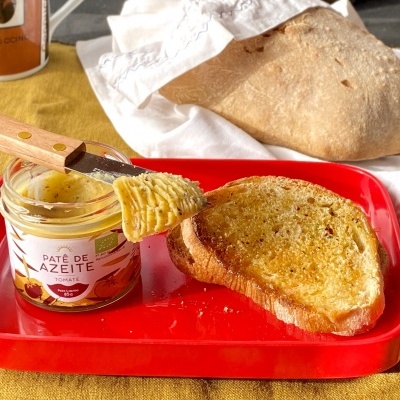 Pão rústico, pão torrado e frasco aberto de paté de azeite com tomate numa tábua vermelha sobre superfície amarela.