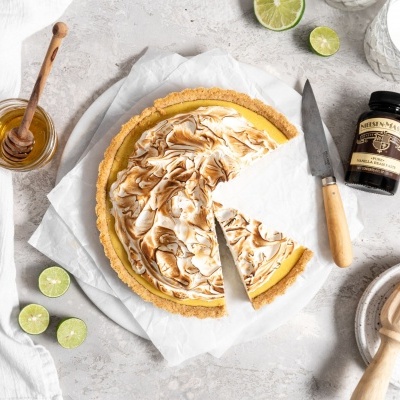 Tarte com merengue dourado, faca, mel, limas e frasco preto numa superfície clara