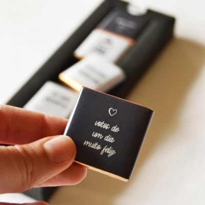 Caixa preta com três chocolates quadrados embalados em papel preto com texto branco