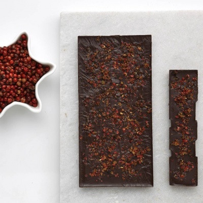 Barra de chocolate escuro com pimenta rosa em superfície branca, com tigela estrela branca cheia de pimenta rosa