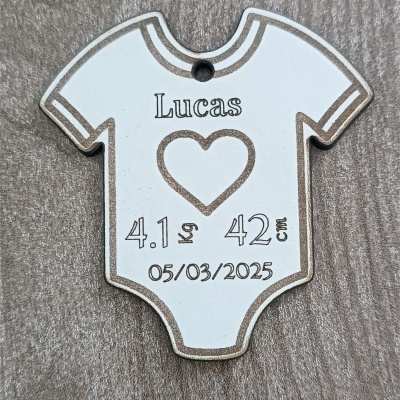 Pingente branco em forma de body de bebé com texto preto e bordas castanhas sobre superfície de madeira clara