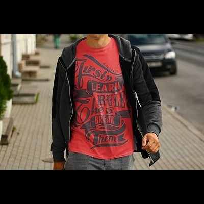 Homem com t-shirt vermelha com texto preto e casaco preto com capuz na rua