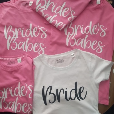 T-shirts rosa e branca com textos Bride's Babes e Bride, sobre superfície de madeira e número 3 branco ao lado.