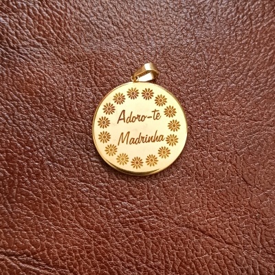 Medalhão dourado redondo com gravação 'Adoro-te Madrinha' e flores, sobre pele castanha.