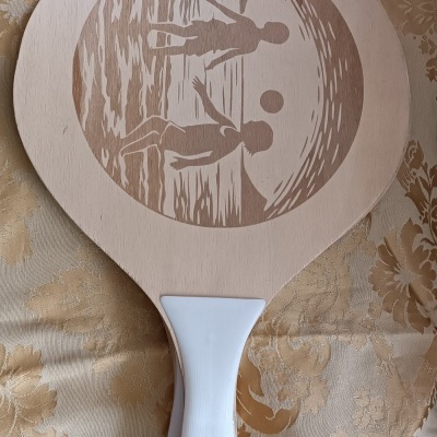 Pá de madeira com gravura de jogadores de voleibol e bola, com pega branca, sobre tecido floral bege.