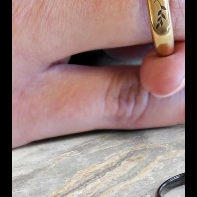 Anel dourado com gravação de folhas e anéis prateados com texto sobre superfície de pedra