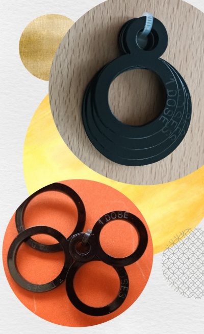 Peças plásticas pretas em anéis interligados com texto '1 DOSE' sobre fundo de madeira e papel colorido.