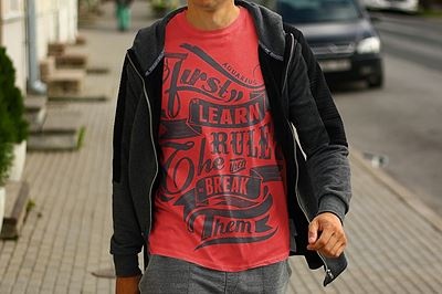 Homem com t-shirt vermelha com texto preto e casaco preto com capuz na rua