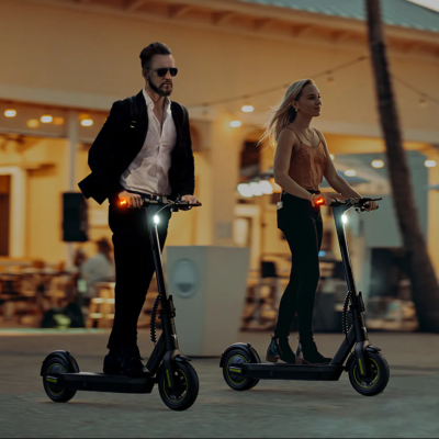 Homem e mulher em trotinetes elétricas pretas com luzes dianteiras ligadas, cenário urbano noturno