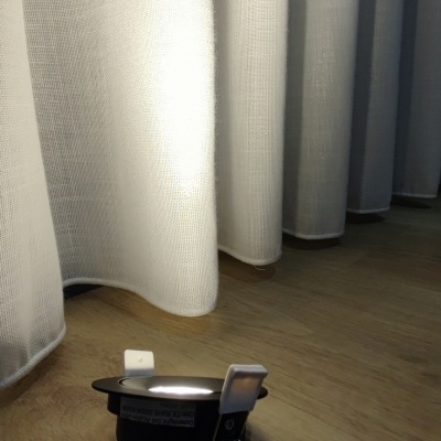 Luz LED embutida redonda preta com cabo branco junto a cortinas brancas no chão de madeira