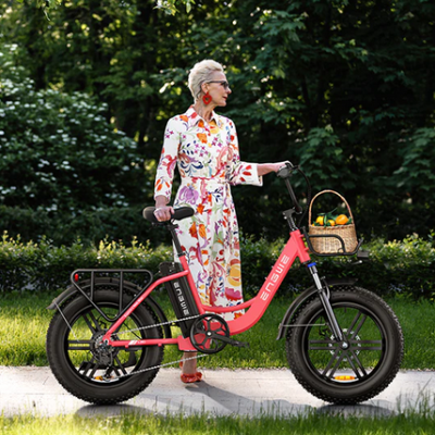 Bicicleta elétrica cor de salmão com pneus grossos e cesto de vime, senhora com vestido estampado junto a relva e árvores