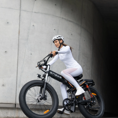 Bicicleta elétrica preta ENGWE com pneus grossos e luz dianteira, mulher de fato branco e capacete a andar em ambiente urbano de betão