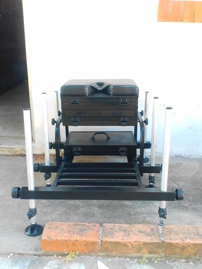 Estrutura metálica preta com almofadas de couro sintético e gavetas, em chão de cimento com tijolos à frente