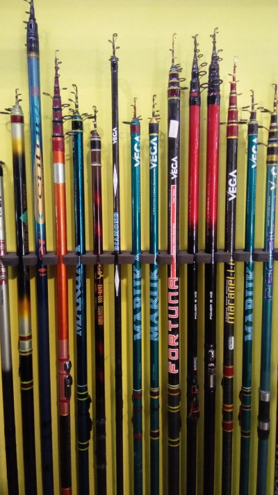 Várias canas de pesca telescópicas coloridas organizadas num suporte em frente a uma parede amarela.