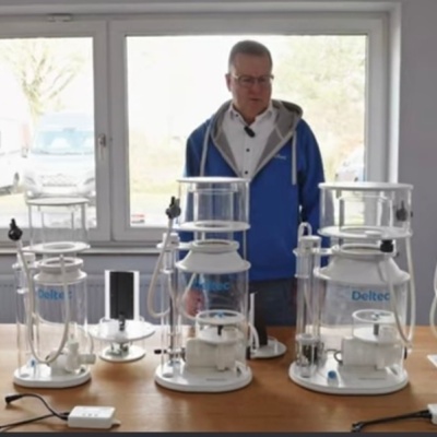 Cinco balões de vidro de vários tamanhos com texto DeReo, sobre mesa em ambiente com janelas ao fundo e homem ao centro.