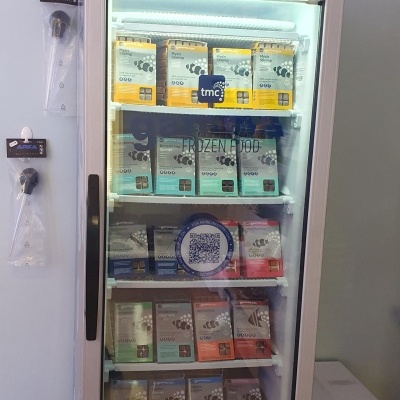 Frigorífico transparente com pacotes coloridos de produtos alimentares em prateleiras e sinalização com preços e descontos em português.