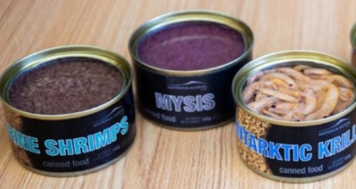 Três latas abertas de comida enlatada para animais com rótulos 'BLUE SHRIMPS', 'MYSIS' e 'ANTARCTIC KRILL' sobre superfície de madeira.