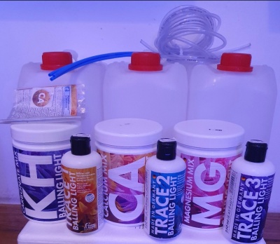 Produtos para aquário em potes e frascos brancos com tampas vermelhas e pretas, rotulados com KH, CA, MG, TRACE 2 e TRACE 3, com tubos ao fundo