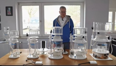 Cinco balões de vidro de vários tamanhos com texto DeReo, sobre mesa em ambiente com janelas ao fundo e homem ao centro.