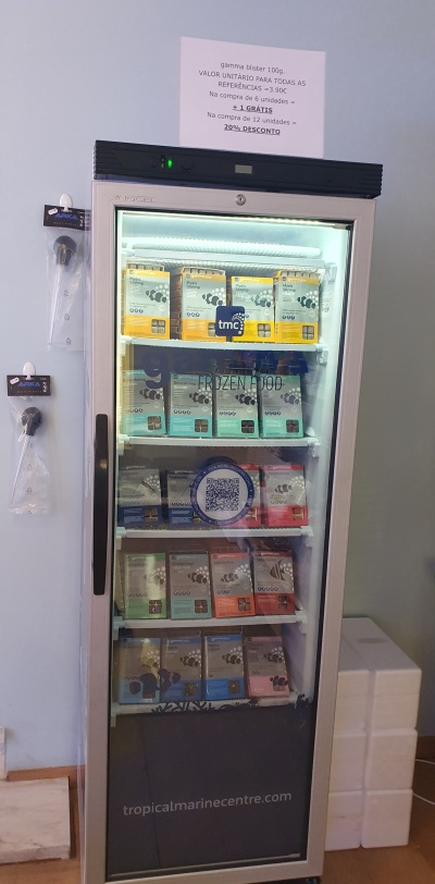 Frigorífico transparente com pacotes coloridos de produtos alimentares em prateleiras e sinalização com preços e descontos em português.