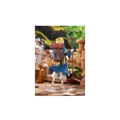 Figura de personagem loira com túnica azul e mochila castanha, segurando um cajado, em suporte transparente, sobre mesa rústica com plantas e objetos decorativos.