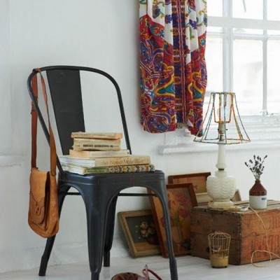 Cadeira preta com livros e mala, sapatos castanhos no chão, mesa com lâmpada e cortina colorida junto a janela