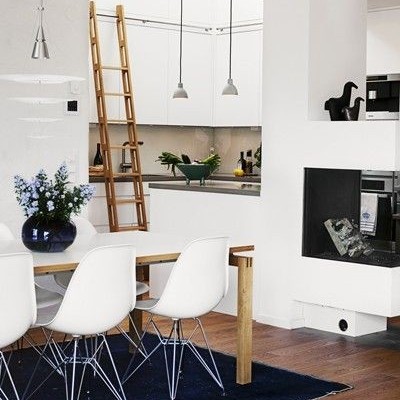 Sala de jantar moderna com mesa de madeira e cadeiras brancas, cozinha branca ao fundo e lareira embutida