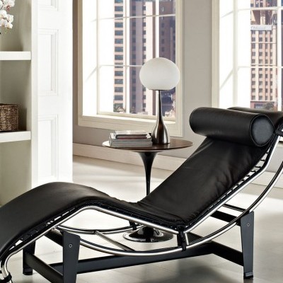 Espreguiçadeira preta em pele com estrutura metálica num interior luminoso próximo de mesa com candeeiro.