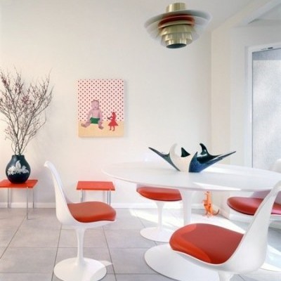 Sala de jantar moderna com mesa branca, cadeiras brancas com estofos vermelhos, vaso azul, quadro com bolinhas vermelhas, e candeeiro pendente.