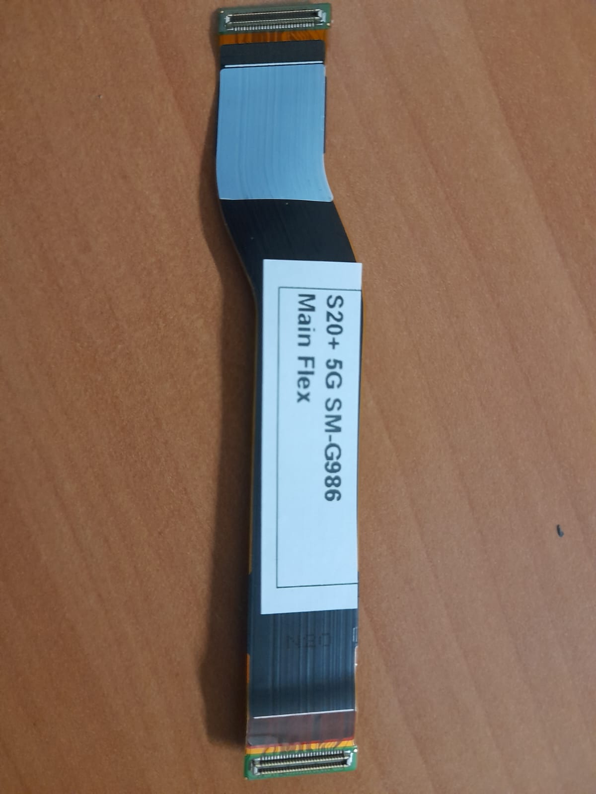 Cabo Flex Principal Samsung Galaxy S20+ (SM-G986) – Original OEM – Testado Cabo flexível preto com conectores dourados e etiqueta branca sobre superfície de madeira clara