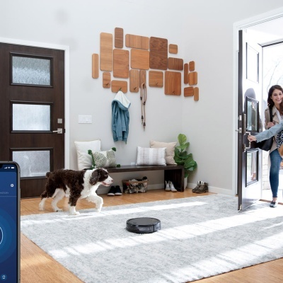 Sala de entrada com porta escura, banco com almofadas, cabideiro de madeira, cão e aspirador robô no chão.