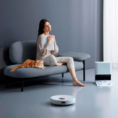 Robô aspirador de pó branco no chão, estação de carga branca, mulher sentada no sofá cinzento com gato laranja