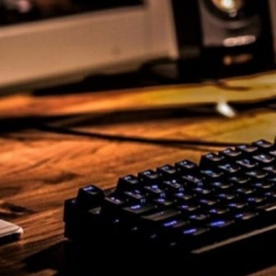 Teclado mecânico preto com luz azul numa secretária de madeira com smartphone branco