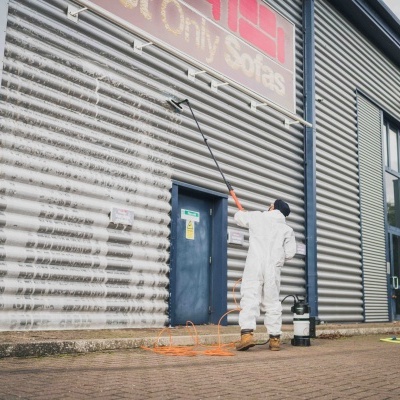 Pessoa com fato protetor a limpar parede metálica com equipamento e lança de limpeza ao ar livre.