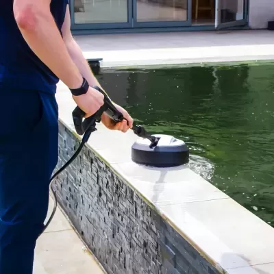Pessoa a limpar bordo de piscina com equipamento elétrico perto de parede de azulejos cinzentos.