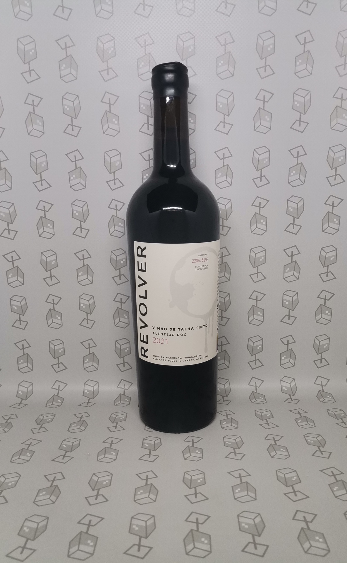 Garrafa de vinho tinto revolver 2021 com rótulo branco sobre fundo cinza com padrão geométrico