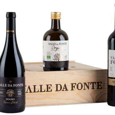 Três produtos Valle da Fonte: duas garrafas de vinho tinto e uma pequena garrafa de azeite à frente de uma caixa de madeira com texto.