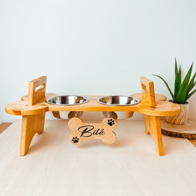 Comedouro duplo para cães em madeira clara com placas personalizadas e tigelas de aço inox, planta ao fundo