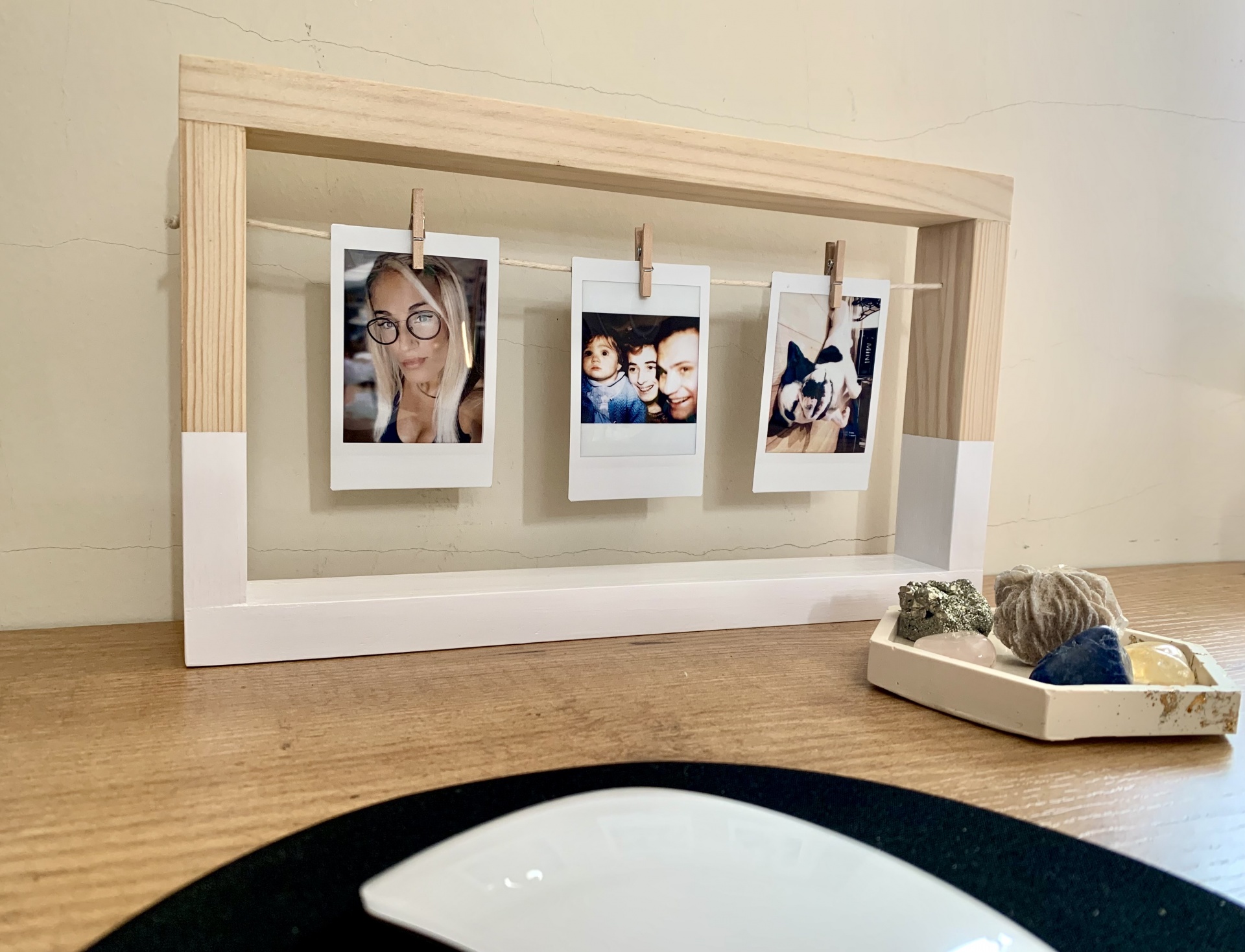 Moldura de madeira com fotos Polaroid penduradas e bolas de fio num tabuleiro sobre superfície de madeira