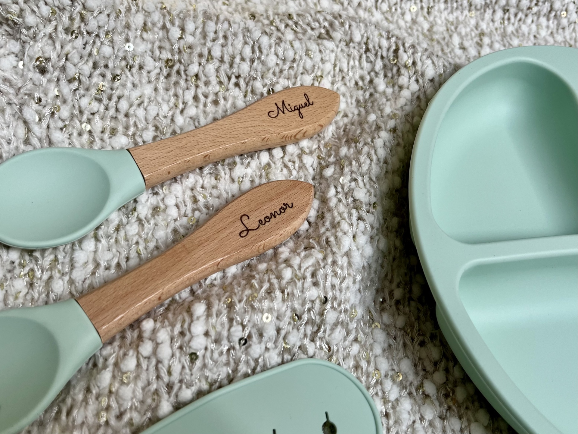 Talheres infantis de silicone verde água com cabos de madeira gravados com os nomes Miguel e Leonor ao lado de prato dividido verde água sobre tecido branco com detalhes dourados