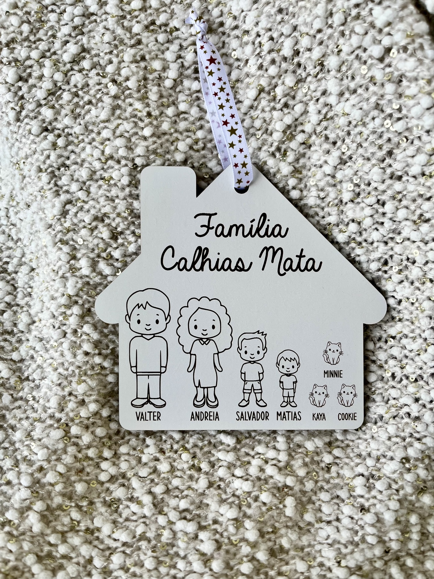 Placa decorativa em forma de casa com ilustrações a preto de uma família e gatos, pendurada por fita com estrelas numa superfície felpuda.