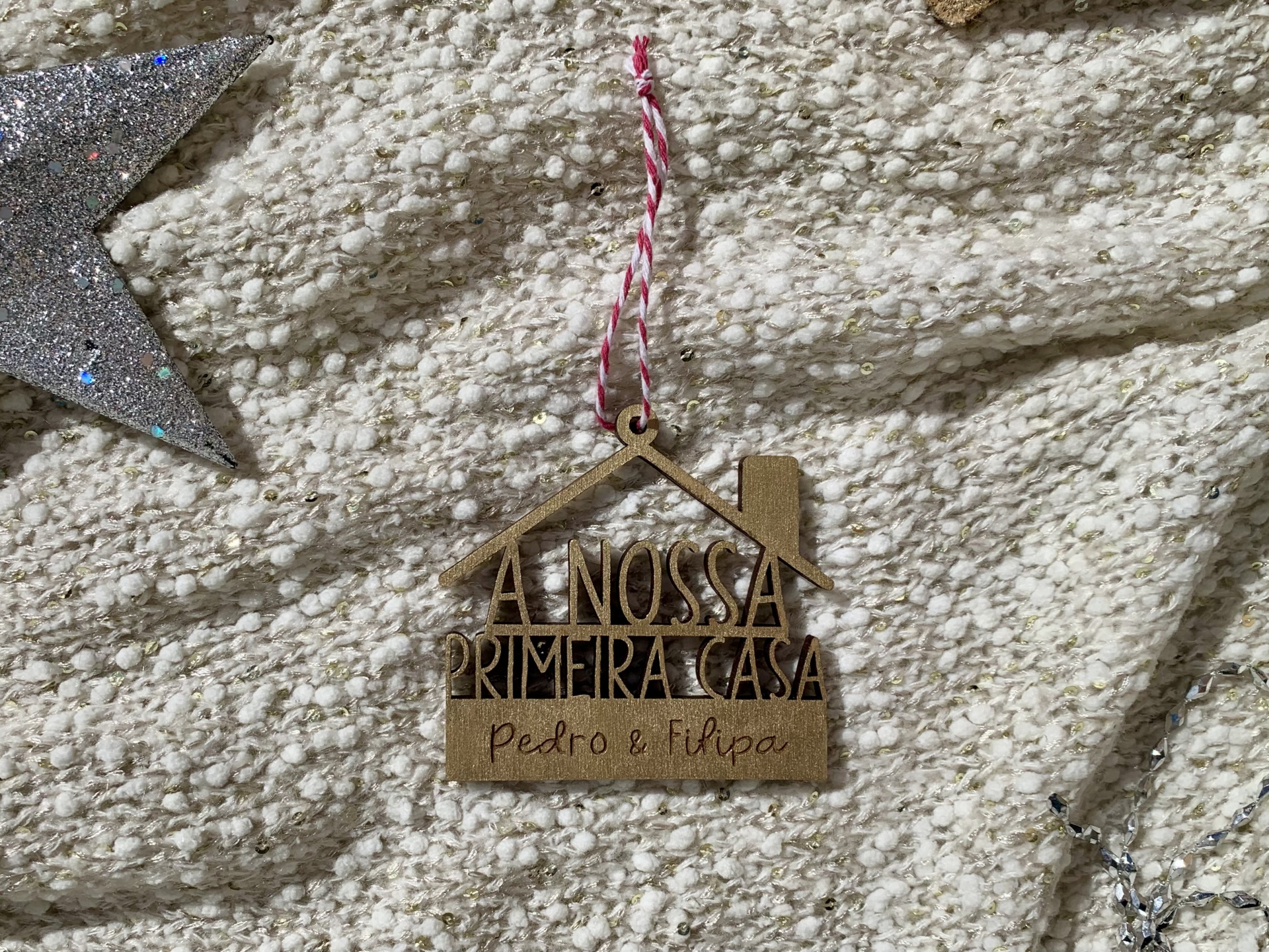 Enfeite pendente de madeira em forma de casa com texto e nomes, sobre tecido bege texturado