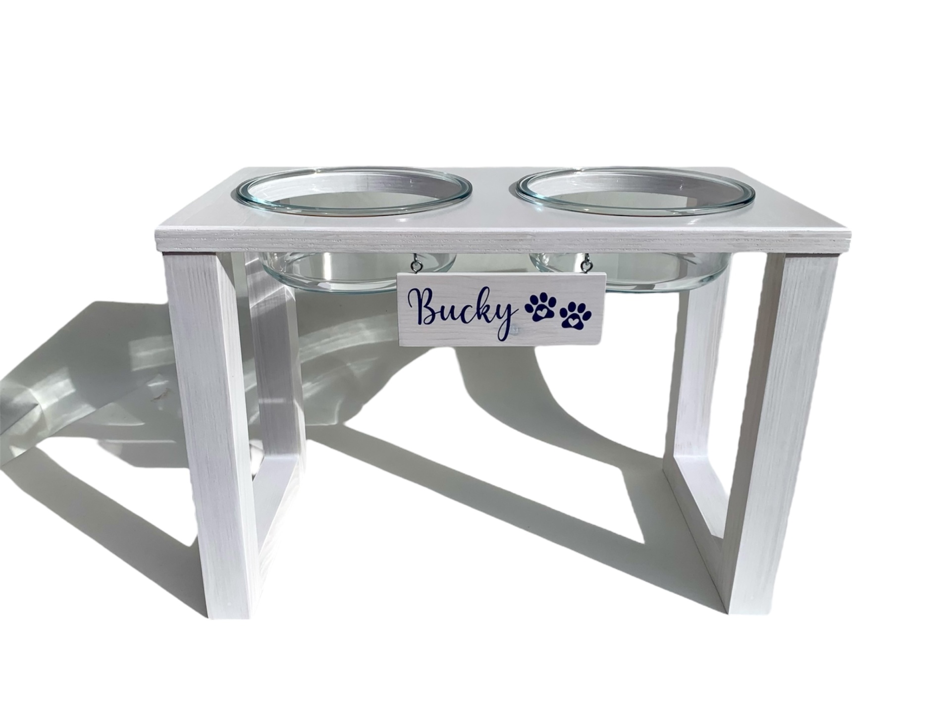 Comedouro elevado branco para animais com duas taças de metal e placa com texto Bucky